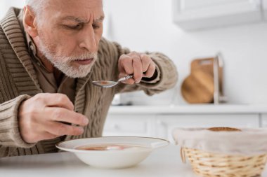 aged man with parkinson disease holding spoon in trembling hand while eating soup in kitchen clipart