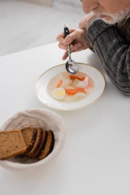 partial view of aged man suffering from parkinson disease and sitting with spoon near soup and bread in kitchen