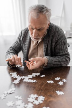 aged man with parkinsonian syndrome and tremor in hands combining jigsaw puzzle on table at home clipart