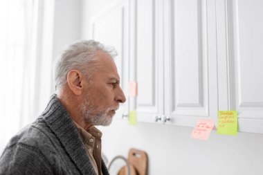 side view of grey haired man with alzheimer syndrome looking at sticky notes with names and phone numbers in kitchen