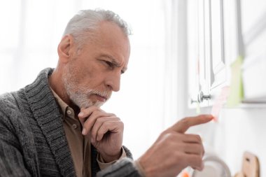 side view of pensive man with alzheimer syndrome touching chin and pointing at blurred sticky note in kitchen
