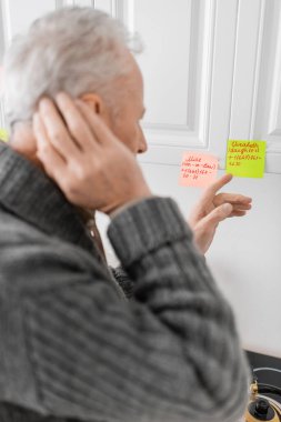 blurred man suffering from memory loss and pointing at sticky notes with names and phone numbers in kitchen