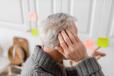 grey haired senior man with alzheimer syndrome touching head and pointing with blurred sticky note in kitchen
