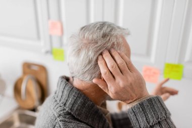 grey haired senior man with alzheimer syndrome touching head and pointing with blurred sticky note in kitchen clipart