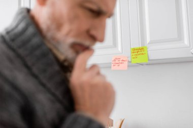 blurred thoughtful man with memory loss touching chin while thinking near sticky notes in kitchen clipart