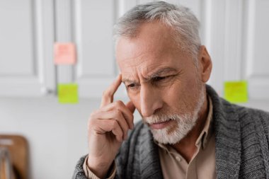 thoughtful senior man with alzheimer disease touching head near blurred sticky notes in kitchen