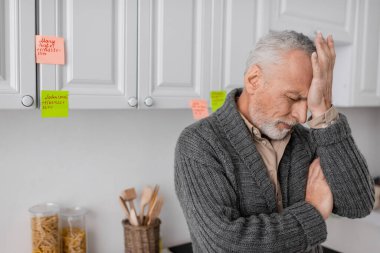 depressed man with alzheimer disease touching forehead while standing near sticky notes in kitchen