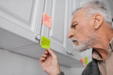 senior man with alzheimer syndrome looking at sticky note with name and phone number in kitchen