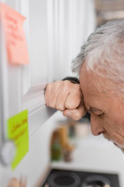 side view of depressed senior man suffering from memory loss while standing near blurred sticky notes in kitchen