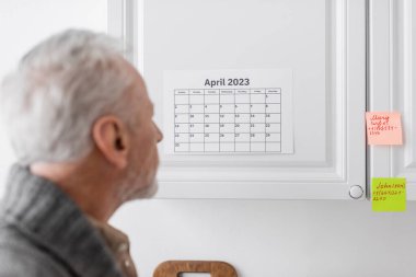 blurred senior man suffering from memory loss and looking at calendar and sticky notes in kitchen