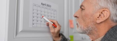 side view of senior man with alzheimer disease pointing with felt pen at calendar in kitchen, banner