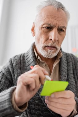 pensive senior man holding felt pen and sticky notes while suffering from memory loss 