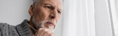 bearded senior man with alzheimer syndrome holding hand near chin and looking away at home, banner