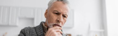 pensive senior man with alzheimer disease holding hand near chin while thinking at home, banner