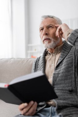 senior man suffering from memory loss and touching head while thinking on couch with blurred notebook