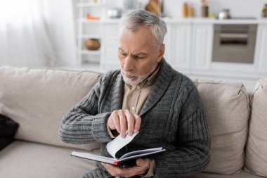 grey haired man suffering from memory loss caused by alzheimer disease looking in notebook on couch at home clipart