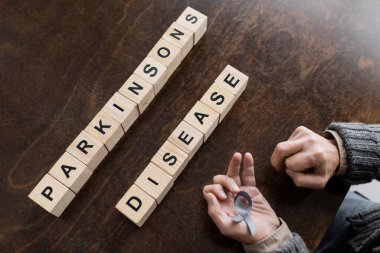 high angle view of cropped man holding grey ribbon in trembling hands cubes with parkinsons disease lettering on wooden table clipart
