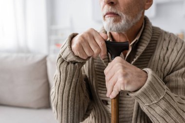 partial view of senior bearded man with parkinson syndrome sitting with walking cane at home