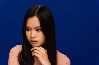 Pensive brunette asian woman looking away isolated on blue 