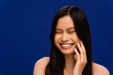 Cheerful asian woman touching face and closing eyes isolated on blue 