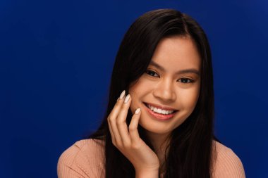 Asian woman with perfect skin touching face isolated on blue 