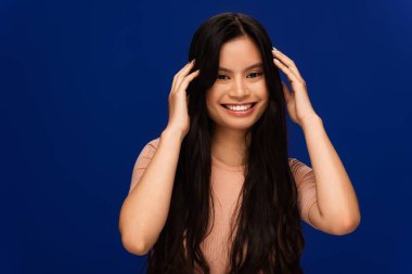 Cheerful asian woman touching long hair isolated on blue  clipart
