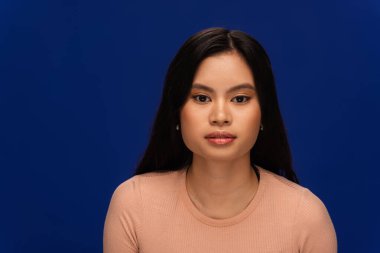 Brunette asian woman in beige t-shirt looking at camera isolated on blue 