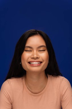 Portrait of joyful asian model closing eyes isolated on blue 