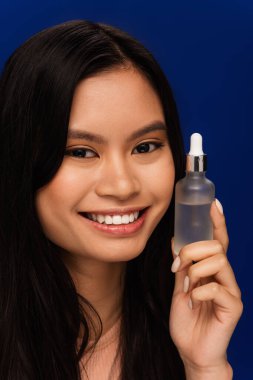Positive asian woman holding serum and looking at camera isolated on blue  clipart