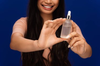 Cropped view of blurred woman holding bottle with cosmetic serum isolated on blue 
