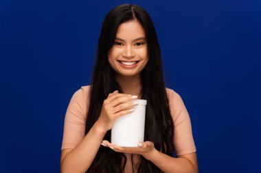 Brunette asian woman holding jar with cosmetic product isolated on blue 