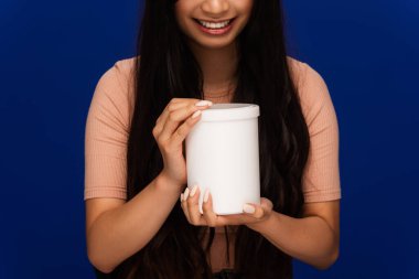 Partial view of positive woman holding jar of cosmetic product isolated on blue  clipart