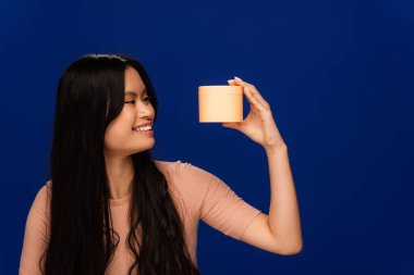 Cheerful asian woman looking at container with hair mask isolated on blue 