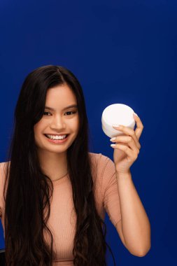 Cheerful long haired asian woman holding container with cream isolated on blue  clipart
