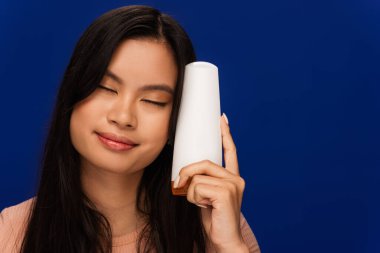 Asian woman with closed eyes holding hair conditioner isolated on blue 