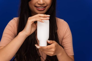 Cropped view of positive brunette woman holding cosmetic lotion isolated on blue 