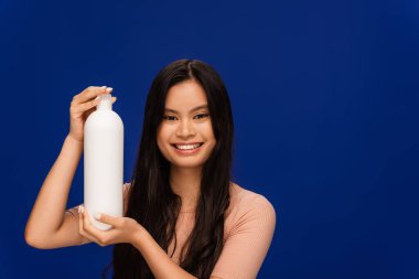 Brunette asian woman holding shampoo and looking at camera isolated on blue  clipart
