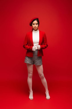 Full length of stylish asian model in beret looking at camera on red background 