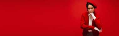 Stylish asian model in beret and white gloves looking at camera isolated on red, banner 
