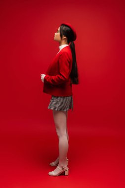 Side view of stylish asian woman in white glove and tights standing on red background 