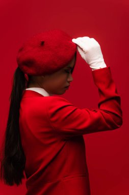 Side view of brunette asian model in blazer touching beret isolated on red 