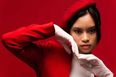 Portrait of asian model in beret and gloves holding hands near face isolated on red 