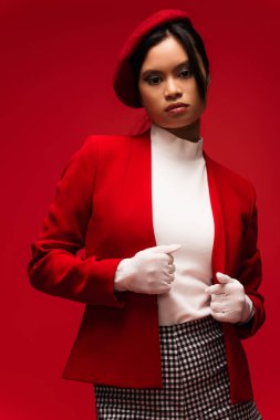 Trendy brunette asian woman in jacket looking at camera isolated on red 