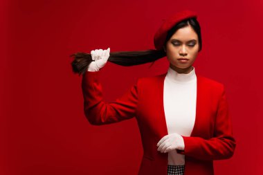 Asian model in gloves and beret touching hair isolated on red 