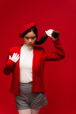 Stylish asian model in beret touching hair while standing isolated on red 