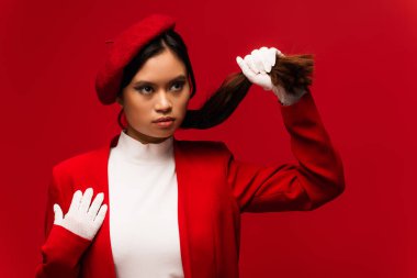Pretty asian woman in beret and jacket touching hair isolated on red 