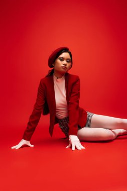 Trendy asian model in jacket and beret sitting on red background and looking at camera 