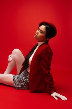 Brunette asian woman in beret and jacket sitting on red background 