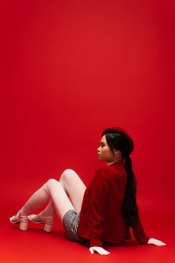 Side view of stylish asian model in white tights and gloves looking away while sitting on red background 