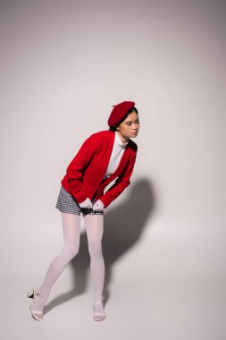 Full length of stylish asian model in beret and checkered shorts posing on grey background 
